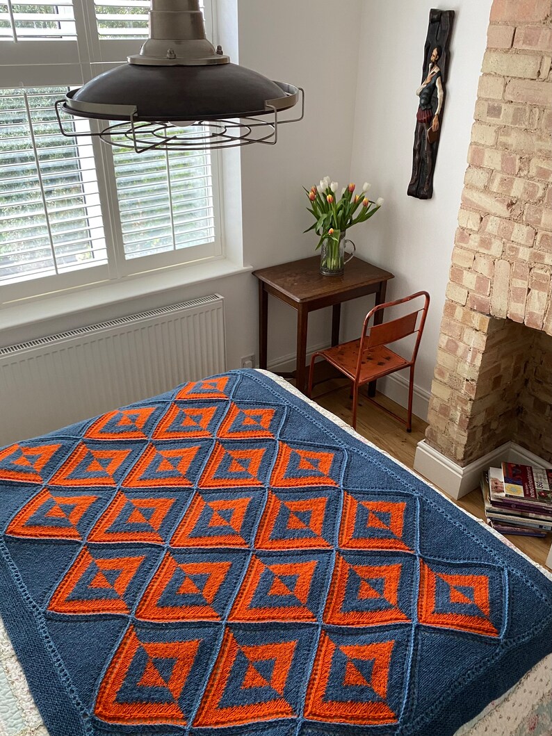 Pode incluir: Uma manta de malha azul e laranja com um padr&atilde;o geom&eacute;trico. A manta est&aacute; em uma cama em um quarto com uma parede branca e uma janela com persianas brancas.