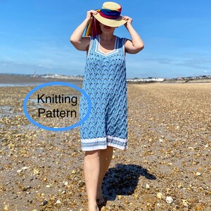 Op de afbeelding: Een vrouw draagt een lichtblauwe gebreide jurk zonder mouwen met een witte rand. Ze staat op een strand met een blauwe lucht op de achtergrond. De jurk heeft een kabelbreipatroon. De tekst "Knitting Pattern" staat in een cirkel op de afbeelding.