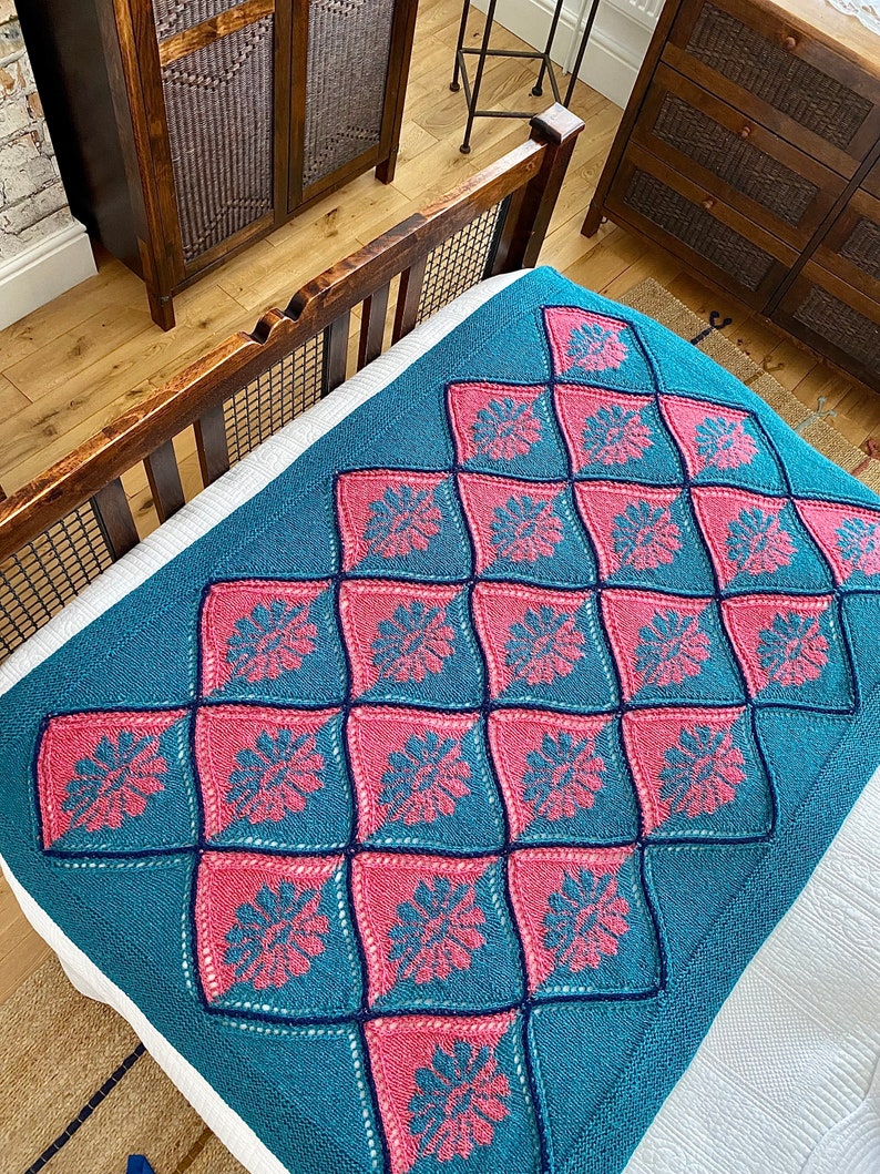 K&ouml;nnte beinhalten: Eine tealfarbene und rosafarbene gestrickte Wolldecke mit einem Rautenmuster. Die Decke ist aus einem strukturierten Garn gefertigt und hat ein zartes Blumenmuster.