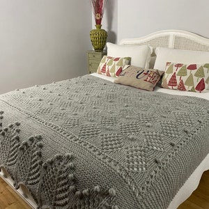 May include: A gray knitted throw blanket with a textured pattern and pom-poms on a white bed with white pillows and a patterned pillow that says "Love".