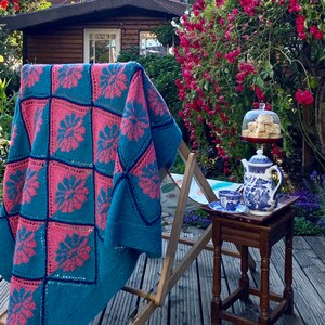K&ouml;nnte beinhalten: Eine teal- und rosafarbene H&auml;keldecke, die &uuml;ber einen Holz-Liegestuhl in einem Garten gelegt wird. Ein kleiner Holztisch mit einer Teekanne und einem Teller mit Geb&auml;ck steht auf der Terrasse.
