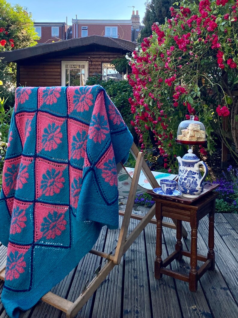 K&ouml;nnte beinhalten: Eine teal- und rosafarbene H&auml;keldecke, die &uuml;ber einen Holz-Liegestuhl in einem Garten gelegt wird. Ein kleiner Holztisch mit einer Teekanne und einem Teller mit Geb&auml;ck steht auf der Terrasse.