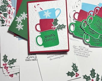 May include: A collection of handmade Christmas greeting cards featuring a stack of three mugs in light blue, red, and green. The green mug displays the text Season's Greetings. The cards include embossed snowflake patterns, holly leaf accents, and printed holiday messages.