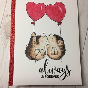 Puede incluir: Una tarjeta de San Valent&iacute;n con dos erizos sosteniendo globos rojos en forma de coraz&oacute;n. Los erizos se besan y la tarjeta dice "siempre & para siempre".