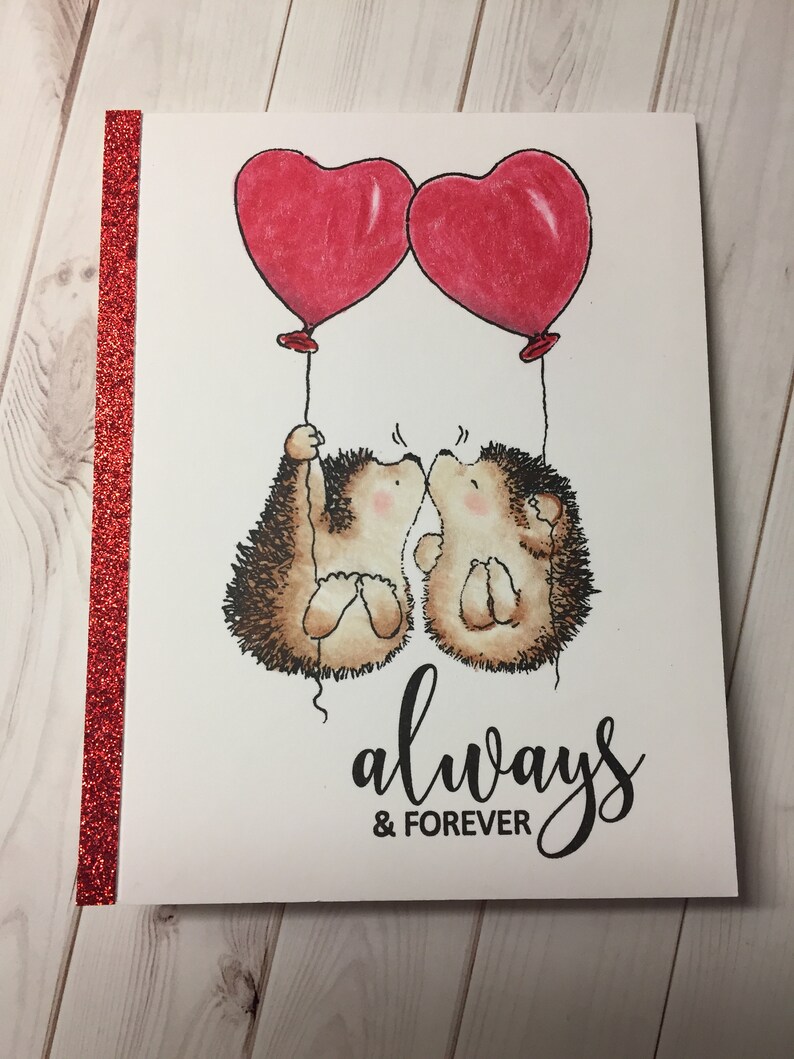 Puede incluir: Una tarjeta de San Valent&iacute;n con dos erizos sosteniendo globos rojos en forma de coraz&oacute;n. Los erizos se besan y la tarjeta dice "siempre & para siempre".