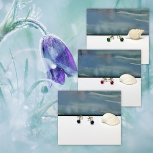 May include: Three pairs of stud earrings with different colored gemstones. The earrings are displayed on a white surface with a seashell next to each pair. The gemstones are green, red, and blue.