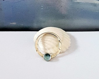 May include: A gold-colored septum ring with a hammered texture and a small, oval, teal-colored gemstone. The ring is displayed on a white seashell against a soft blue and white background.