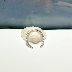 K&ouml;nnte beinhalten: Ein silberner Ring mit geh&auml;mmerter Oberfl&auml;che, in Form eines Halbmondes. Der Ring liegt auf einer wei&szlig;en Muschel.
