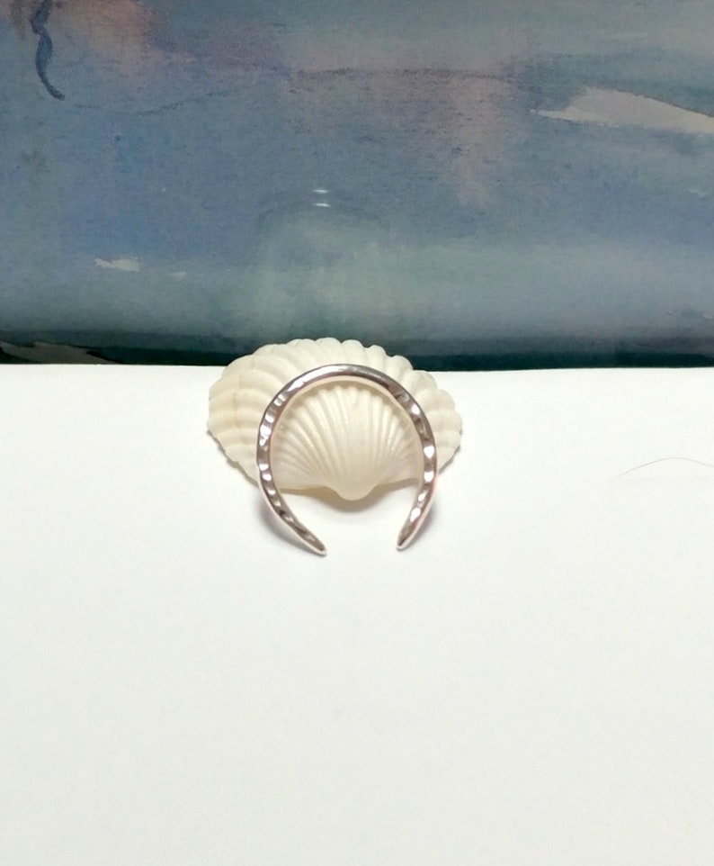 K&ouml;nnte beinhalten: Ein silberner Ring mit geh&auml;mmerter Oberfl&auml;che, in Form eines Halbmondes. Der Ring liegt auf einer wei&szlig;en Muschel.