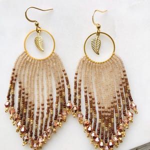 Op de afbeelding: Een paar kralen oorbellen met een goudkleurige bladbedel en een cirkelvormige goudkleurige ring. De oorbellen hebben een franje van lichtbruine, bruine en roze kralen. De oorbellen worden op een wit oppervlak gepresenteerd.