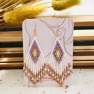 Op de afbeelding: Een paar witte en paarse kralen oorbellen met gouden accenten. De oorbellen worden getoond op een kaart met de tekst "Jonk Beaded Jewels".