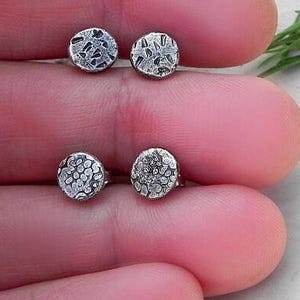 Pode incluir: Quatro pequenos brincos de prata com superfícies texturizadas são exibidos. Dois pares de brincos são visíveis, cada um com um padrão diferente. Os brincos são segurados na mão de uma pessoa, com um fundo branco e um raminho verde no canto superior direito.