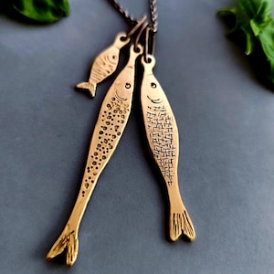 May include: A brass-toned necklace featuring three fish pendants of varying sizes. The fish pendants have etched details, including scales and fins. The necklace is displayed on a gray surface, with green leaves in the background.