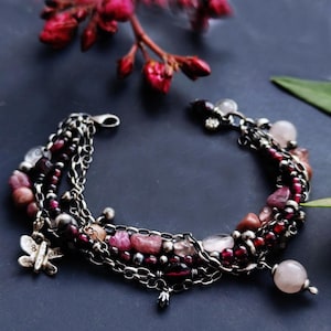 Könnte beinhalten: Ein mehrsträngiges Armband mit dunkel silberfarbenen Metallketten, durchsetzt mit tiefroten Granatperlen, blassrosa Rosenquarzsteinen und kleinen Metallkugeln. Ein silberfarbener Schmetterlingsanhänger hängt an einer Seite des Armbands.