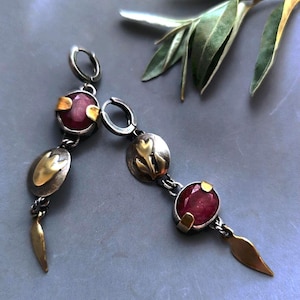Puede incluir: Pendientes plateados con piedras de color rubí y detalles dorados. Los pendientes tienen un diseño colgante con elementos ovalados y en forma de hoja. Se muestran sobre una superficie gris con hojas verdes en el fondo.