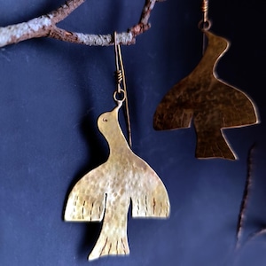 Peut inclure: Une paire de boucles d'oreilles en forme d'oiseau de couleur laiton avec une texture martelée. Les boucles d'oreilles sont suspendues à de fins fils dorés et sont suspendues sur un fond bleu foncé. Les oiseaux ont les ailes déployées et des plumes de queue détaillées.