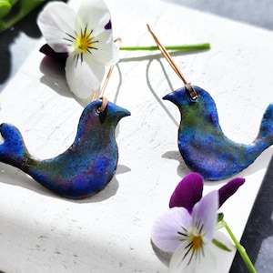 May include: A pair of handcrafted bird-shaped earrings with a blue and green mottled finish. The earrings are displayed with white and purple pansies, showcasing the jewelry's artistic design and color palette.