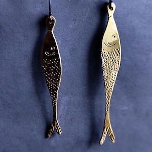 Pode incluir: Um par de brincos dourados em forma de peixe. Cada brinco tem um corpo texturizado, um rosto sorridente e uma pequena cauda. Os brincos penduram-se em fios de orelha pretos. O fundo é azul escuro.