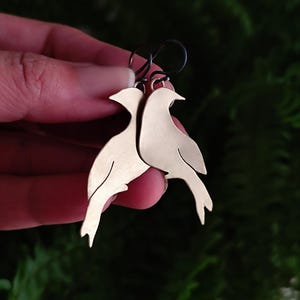 Op de afbeelding: Een paar goudkleurige oorbellen in de vorm van vogels. De oorbellen zijn gemaakt van glanzend metaal en hebben een eenvoudig, elegant ontwerp. De vogels kijken elkaar aan en de oorbellen hangen aan zwarte oorhaken. De achtergrond is vaag groen.