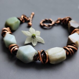 Pode incluir: Uma pulseira artesanal com contas geométricas verde claro e branco sujo, conectadas por espirais de arame de cobre. A pulseira é presa com um fecho de alternância de cobre. Uma flor branca com centro amarelo está por perto.