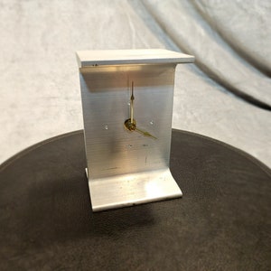 May include: A silver metal clock with gold hands. The clock is shaped like a rectangle with a curved top and bottom.