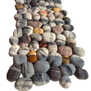 May include: A long, rectangular rug made of felted wool, shaped like a collection of smooth, rounded stones. The stones are in shades of grey, brown, and white, with some yellow and orange accents.