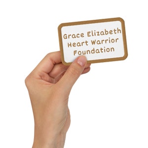 May include: A rectangular white card with rounded corners and a brown border is held in a hand. The card displays the text "Grace Elizabeth Heart Warrior Foundation" in brown letters.