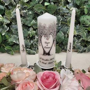 Puede incluir: Dos velas de unidad de boda blancas con caligrafía negra. La vela más grande tiene un diseño de dos árboles formando un corazón con los nombres "Susie & Emmet" y la fecha "6 de octubre de 2025" y la ubicación "St Mary's Church".