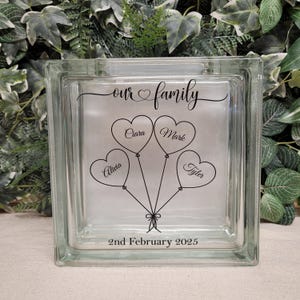 Può includere: Un blocco di vetro con un design di quattro palloncini a forma di cuore con i nomi "Ciara", "Mark", "Allivia" e "Tyler" scritti al loro interno. Il testo "our family" e "2nd February 2025" sono anche inclusi nel design.