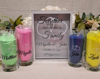 Puede incluir: Un juego de ceremonia de boda con arena de colores. Incluye cinco recipientes de vidrio llenos de arena verde, rosa, azul y amarilla. Un marco blanco dice "Together we make a Family" con nombres y una fecha.