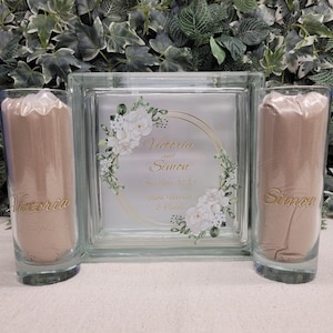 May include: Two clear glass cylinders filled with sand, labeled "Victoria" and "Simon", sit on either side of a square glass vase with a floral design and the text "Victoria and Simon, 2nd July 2023, Faith, Hope & Charity".