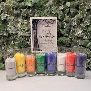 Può includere: Otto barattoli di vetro trasparente riempiti di sabbia colorata, ognuno con un nome scritto sul davanti. I barattoli sono disposti in fila su un tavolo. Il testo sul cartello dietro i barattoli dice "Insieme formiamo una famiglia Papà, Mamma Tiarnan & Rian Cerimonia di denominazione".