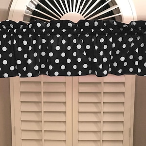 May include: Black and white polka dot valance with a ruffled edge. The valance is hanging on a window with white blinds.