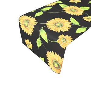 Op de afbeelding: Zwarte tafelloper met een herhaald patroon van gele zonnebloemen en groene bladeren. De zonnebloemen hebben een gedetailleerd ontwerp, met een donker centrum en heldergele bloemblaadjes. De loper is gemaakt van een zachte, duurzame stof.