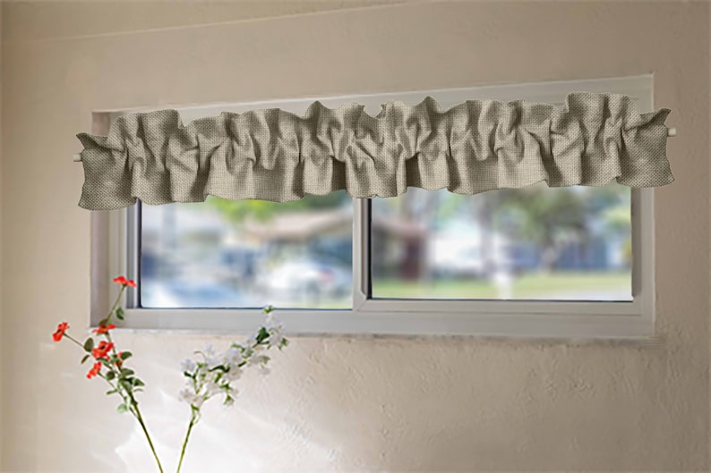 May include: A beige ruffled valance window curtain with a subtle woven texture. The valance is hung on a white window frame with a blurred view of greenery outside.