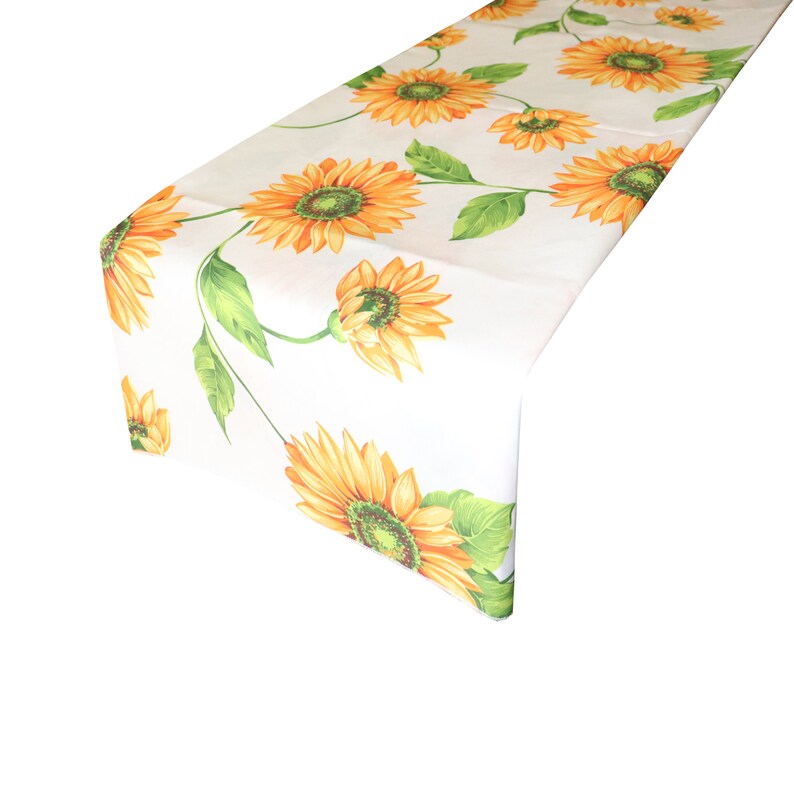 Op de afbeelding: Witte tafelloper met een herhalend patroon van grote zonnebloemen. De zonnebloemen hebben gele bloemblaadjes en groene centra, verbonden door groene stelen en bladeren. De loper is gemaakt van een lichtgekleurde stof.