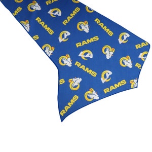 May include: Blue table runner with a repeating pattern of the Los Angeles Rams logo and the word "RAMS" in white and yellow. The runner has a pointed end and is made of fabric.