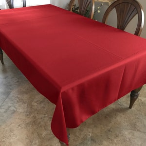May include: A solid red rectangular tablecloth draped over a wooden table. The tablecloth is smooth and wrinkle-free.