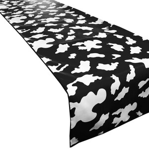 May include: Black and white cow print table runner. The runner is made of a satin-like fabric and has a subtle sheen.