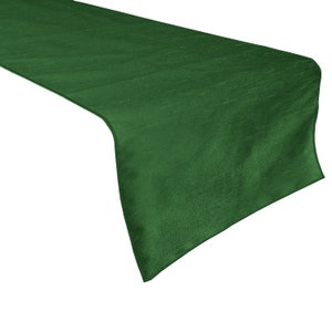 May include: A deep emerald green table runner with a subtle sheen. The runner is made of a smooth, possibly silk-like fabric, and is designed to be placed on a table. The fabric has a slight texture, adding visual interest to the solid color.