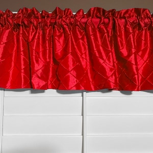 May include: A vibrant red valance with a diamond-stitched pattern. The fabric is gathered at the top, creating a ruffled effect. The valance hangs above white shutters, adding a pop of color to the room.