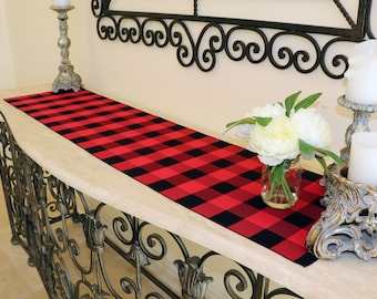 May include: Red and black buffalo plaid table runner on a wooden table with a vase of white flowers and candlesticks.