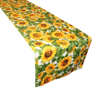 Op de afbeelding: Een lange, rechthoekige tafelloper met een herhaald patroon van gele zonnebloemen met bruine centra en groene bladeren op een witte achtergrond. Het bloemmotief bedekt het gehele oppervlak van de loper.