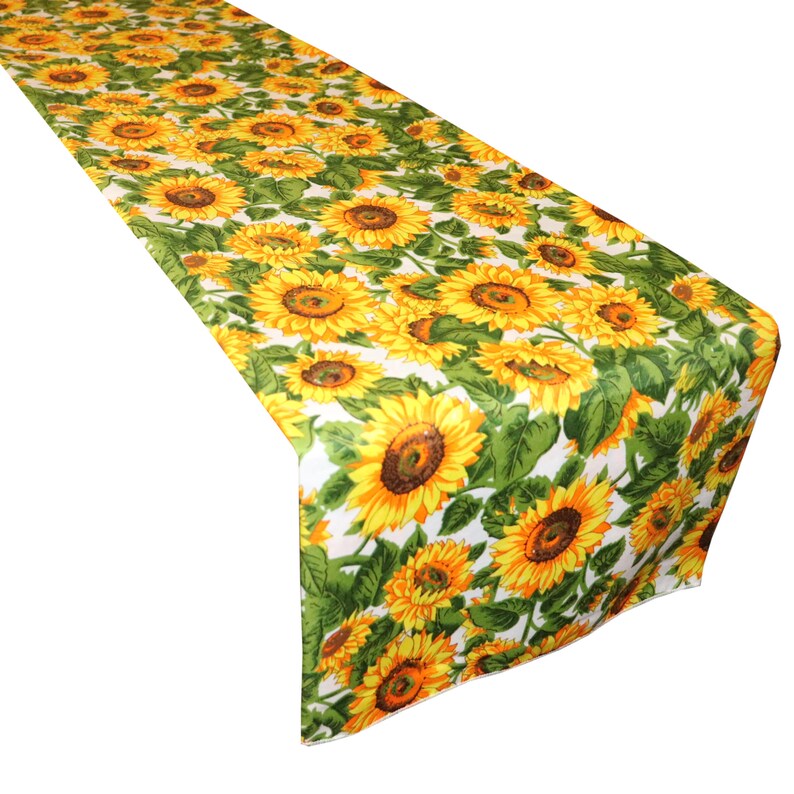 Op de afbeelding: Een lange, rechthoekige tafelloper met een herhaald patroon van gele zonnebloemen met bruine centra en groene bladeren op een witte achtergrond. Het bloemmotief bedekt het gehele oppervlak van de loper.