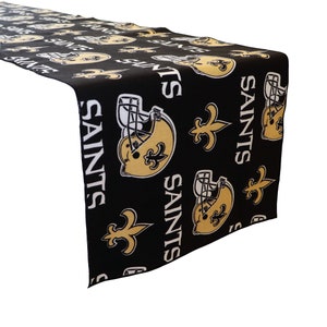 May include: Black and gold table runner featuring a repeating pattern of New Orleans Saints logos, including the team name, fleur-de-lis, and football helmets.