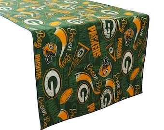 May include: Green and gold table runner with a repeating pattern of Green Bay Packers logos, helmets, and the words "Green Bay Packers".