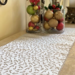 Puede incluir: Un camino de mesa blanco con un patrón repetido de bastones de caramelo beige. Dos jarrones de vidrio llenos de adornos navideños rojos, dorados y verdes se encuentran en el fondo, creando una escena festiva.