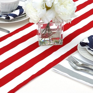 May include: Red and white striped table runner with a clear glass vase filled with white flowers. The table runner is on a white table with a gray and white striped napkin, a white plate, and a silver fork.