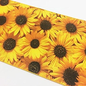 Op de afbeelding: Een close-up van een stof met een zonnebloemmotief. Het ontwerp toont talrijke zonnebloemen met gele bloemblaadjes en donkerbruine centra. De bloemen zijn dicht op elkaar geplaatst, wat een levendige en zomerse uitstraling cre&euml;ert. De achtergrond is wit.