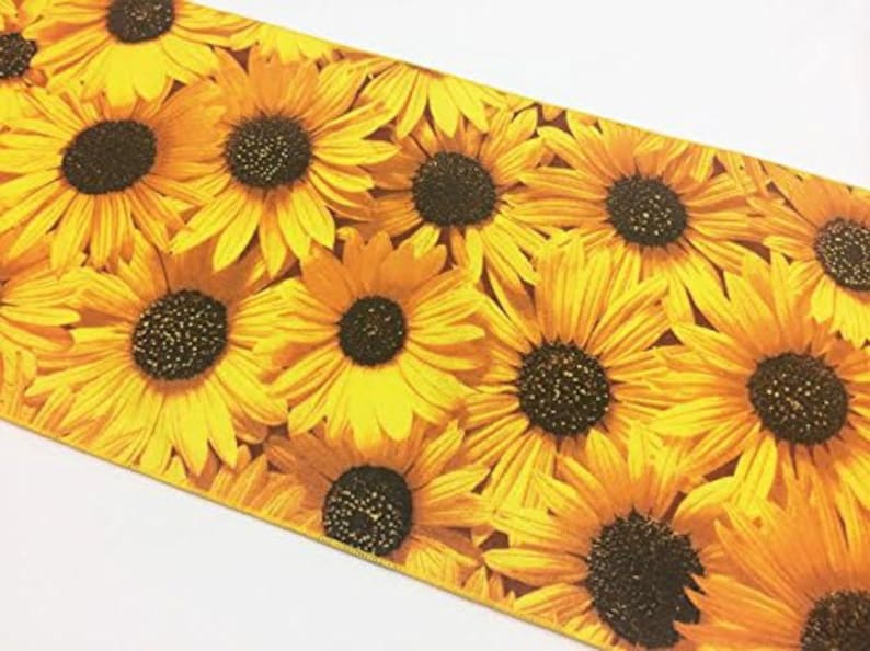 Op de afbeelding: Een close-up van een stof met een zonnebloemmotief. Het ontwerp toont talrijke zonnebloemen met gele bloemblaadjes en donkerbruine centra. De bloemen zijn dicht op elkaar geplaatst, wat een levendige en zomerse uitstraling cre&euml;ert. De achtergrond is wit.